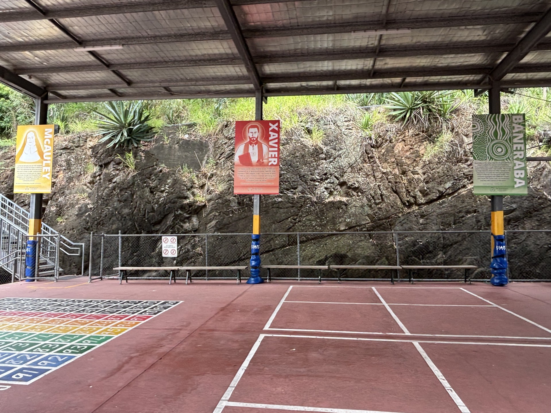 Covered outdoor school play area with painted court markings and Ignatian tradition posters along a rock wall.