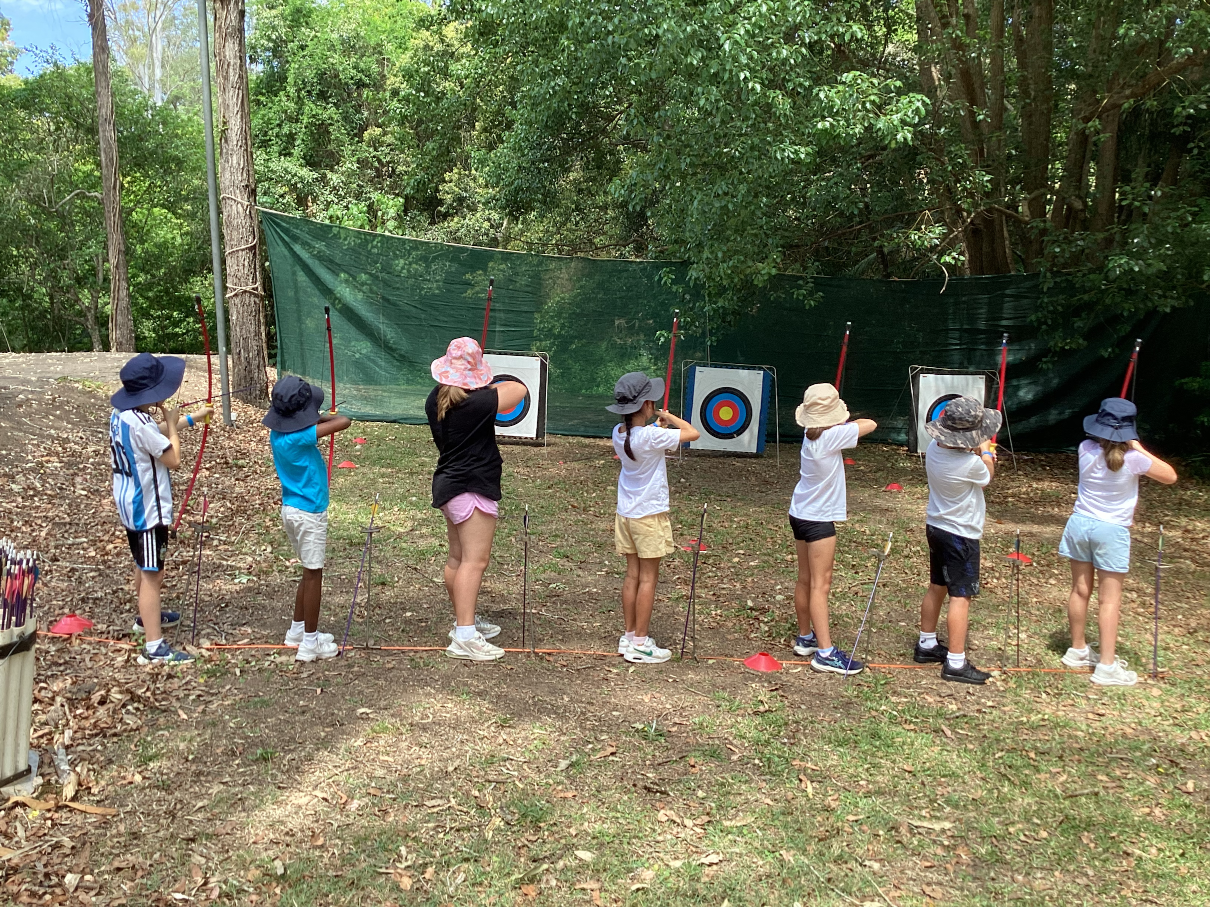 Students lined up outdoors practicing archery and aiming bows at targets during a school activity.