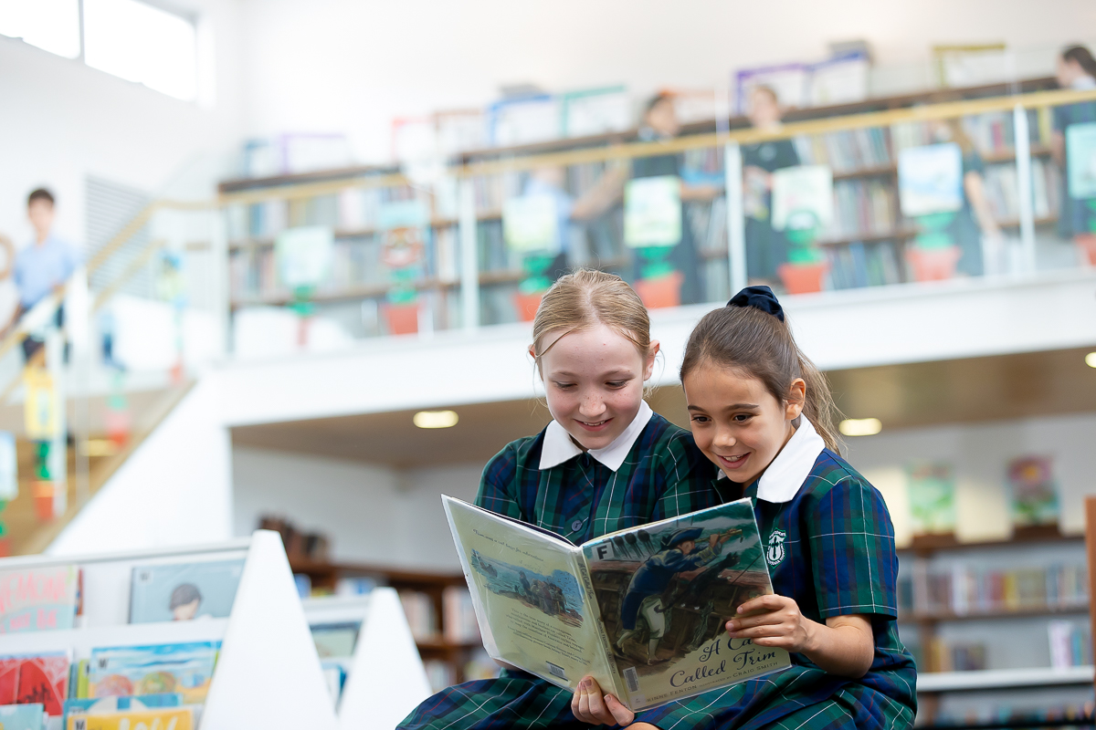 Two students in school uniform reading a book together in a school library.