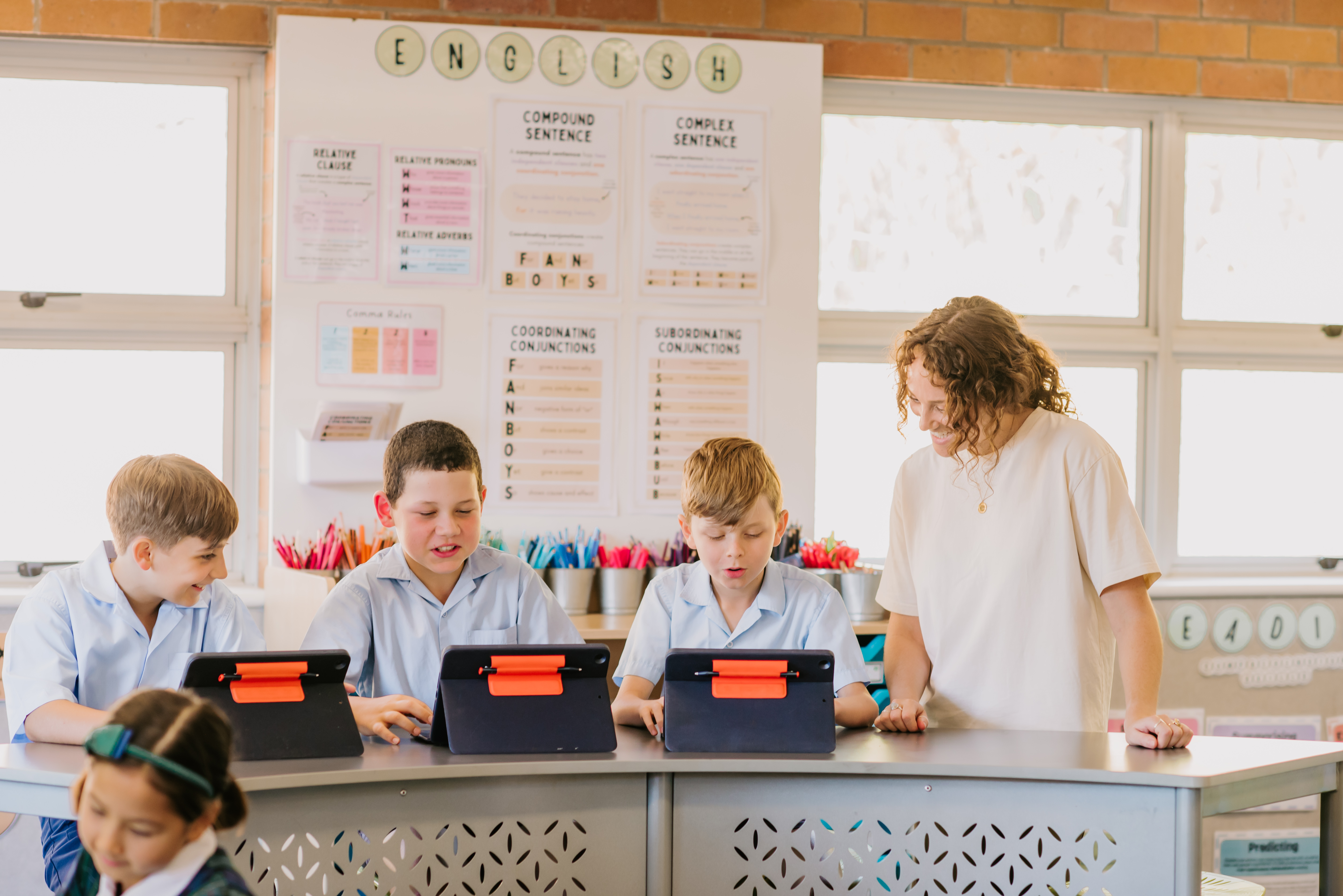 Students using tablets at desks while an adult supports their learning in a classroom.